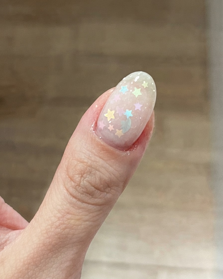 Eine Nahaufnahme eines Daumennagels mit einer milchig-rosa Basis und darauf platzierten bunten Pastell-Stern-Glitzern.