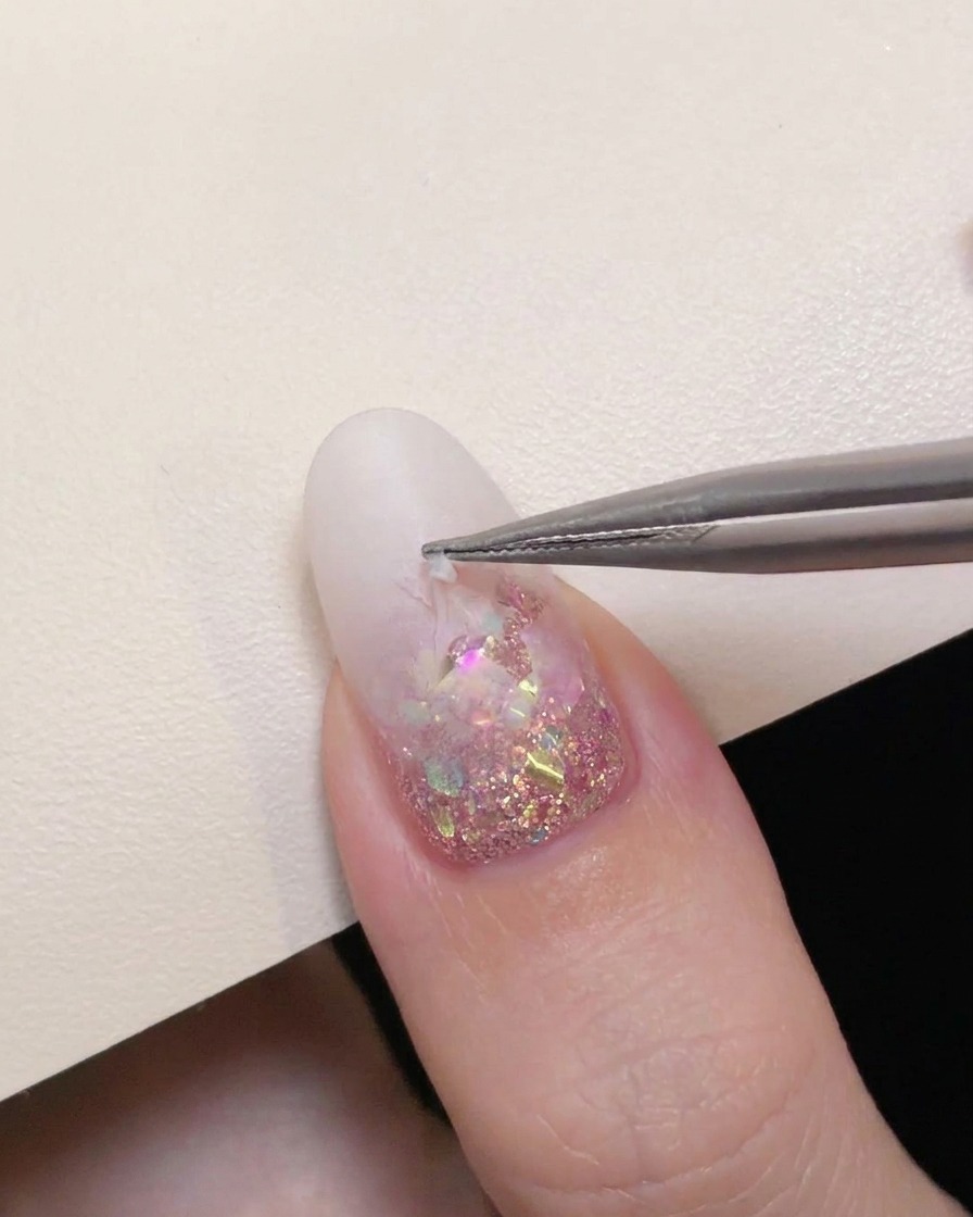 Eine Präzisionspinzette zieht vorsichtig einen hohlen Sternaufkleber von einem milchig weißen Verlaufnagel ab, um den Pink-Glitter darunter freizulegen.