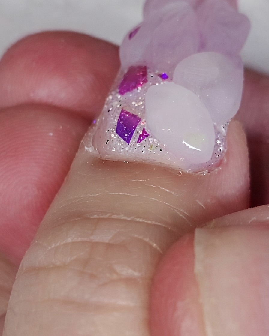Ein Nagelpinsel platziert eine pinke Acrylperle auf einer Glitzer-Nagelbasis, um eine Herzform zu formen.