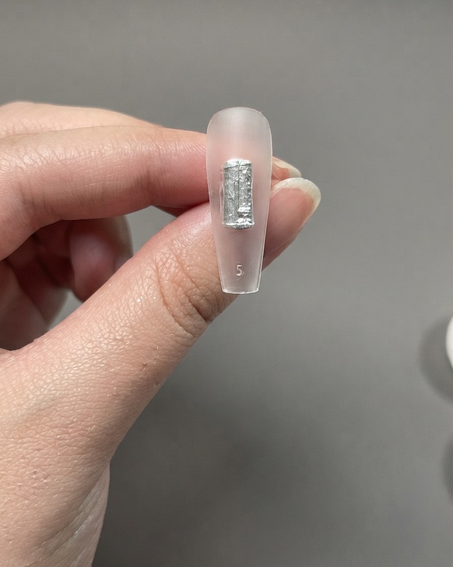 Eine klare Nagelverlängerung zwischen den Fingern, die einen direkt aufgetragenen Pinselstrich ohne Unterlack zeigt.