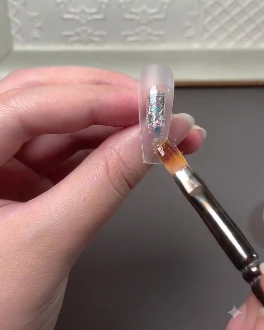 Ein Nailart-Pinsel trägt bunte Glitzerflocken und Hochglanzpuder auf die Mitte einer klaren Nagelverlängerung auf.