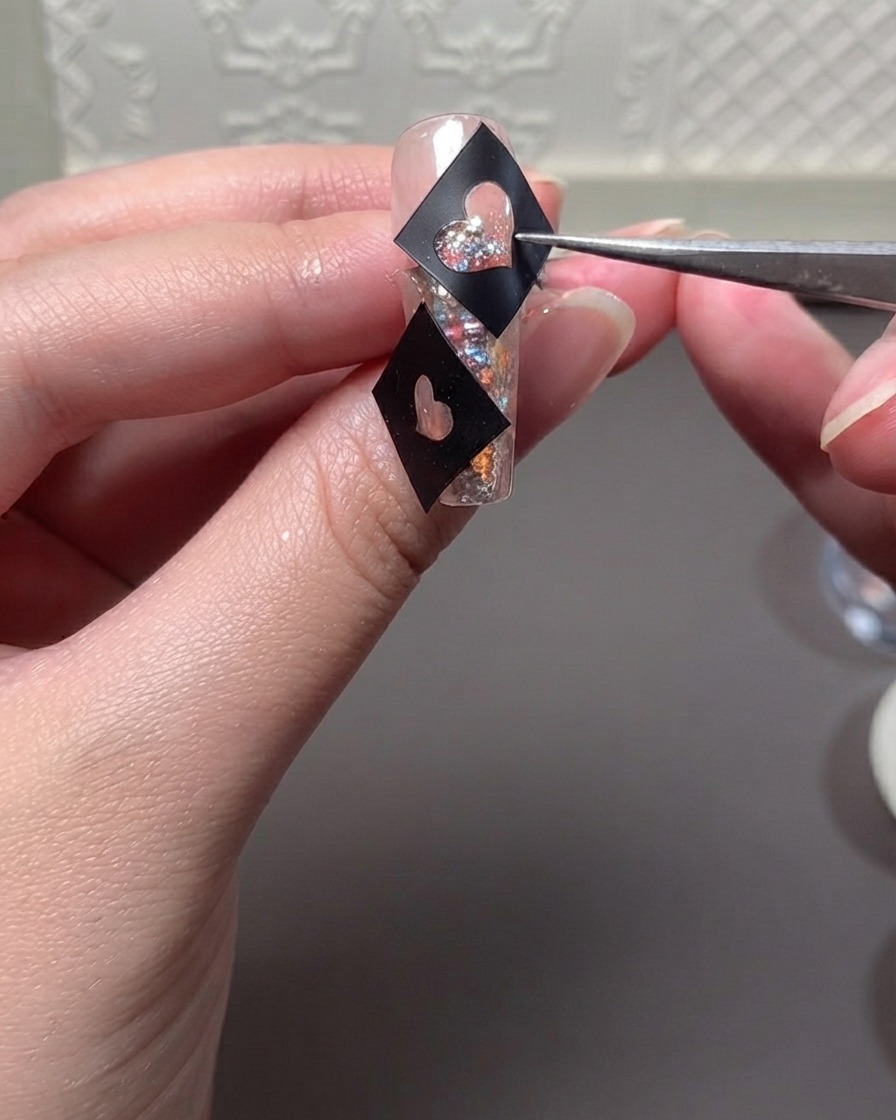 Eine Pinzette hält eine schwarze Herzschablone über einen glitzernden Nagel, während Gel aufgetragen wird.