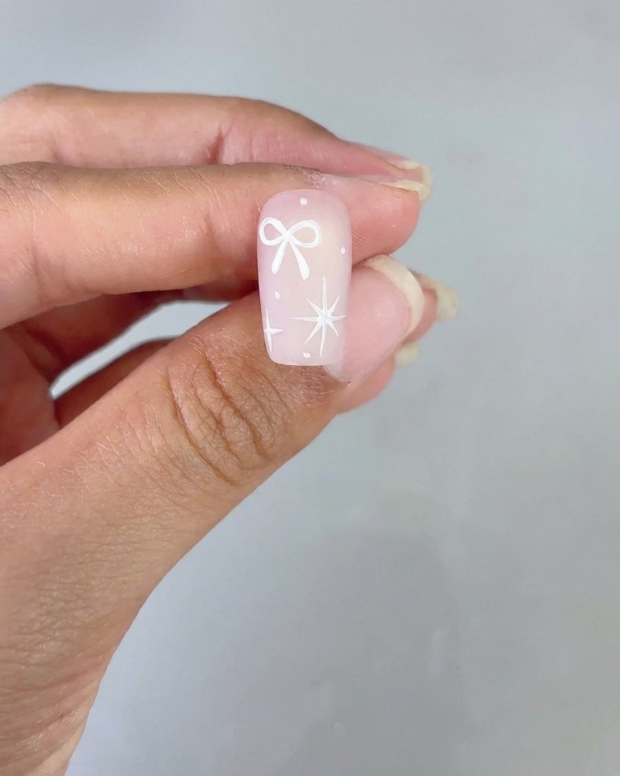Ein fertiges Nageldesign mit einer weißen Schleife und einem vierzackigen Stern auf einer rosa Farbverlauf-Basis wird zur Ansicht hochgehalten.