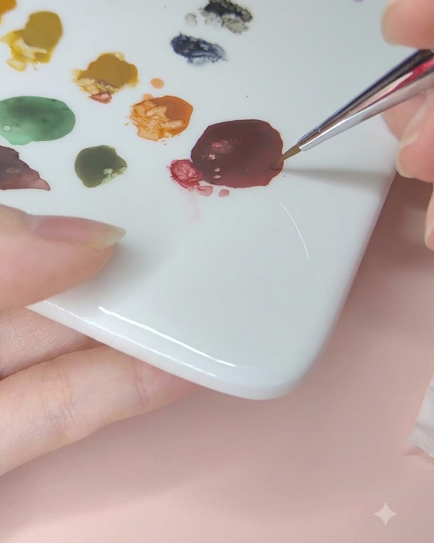 Ein feiner Nailart-Pinsel nimmt dunkelrote Aquarellfarbe von einer Keramikpalette auf.