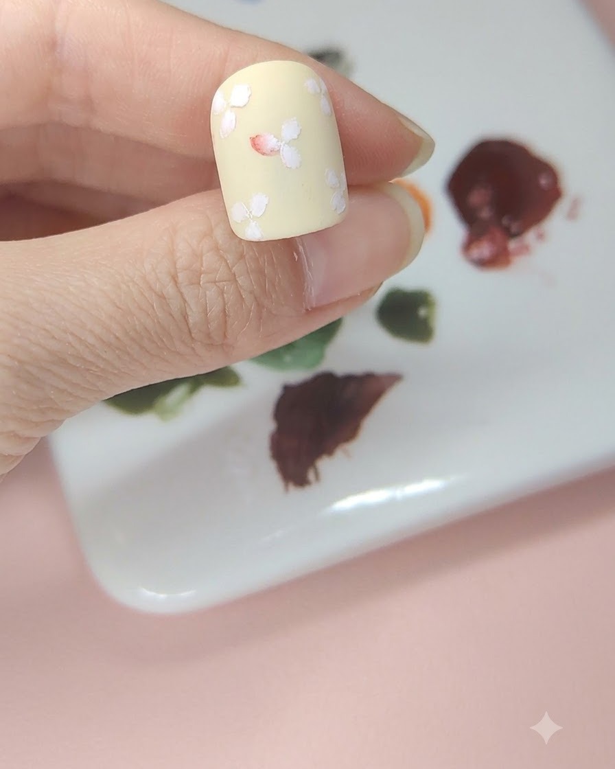 Ein blassgelber Nagel mit weißen Blumen, bei dem die Mitte der mittleren Blume mit roter Aquarellfarbe getönt ist.