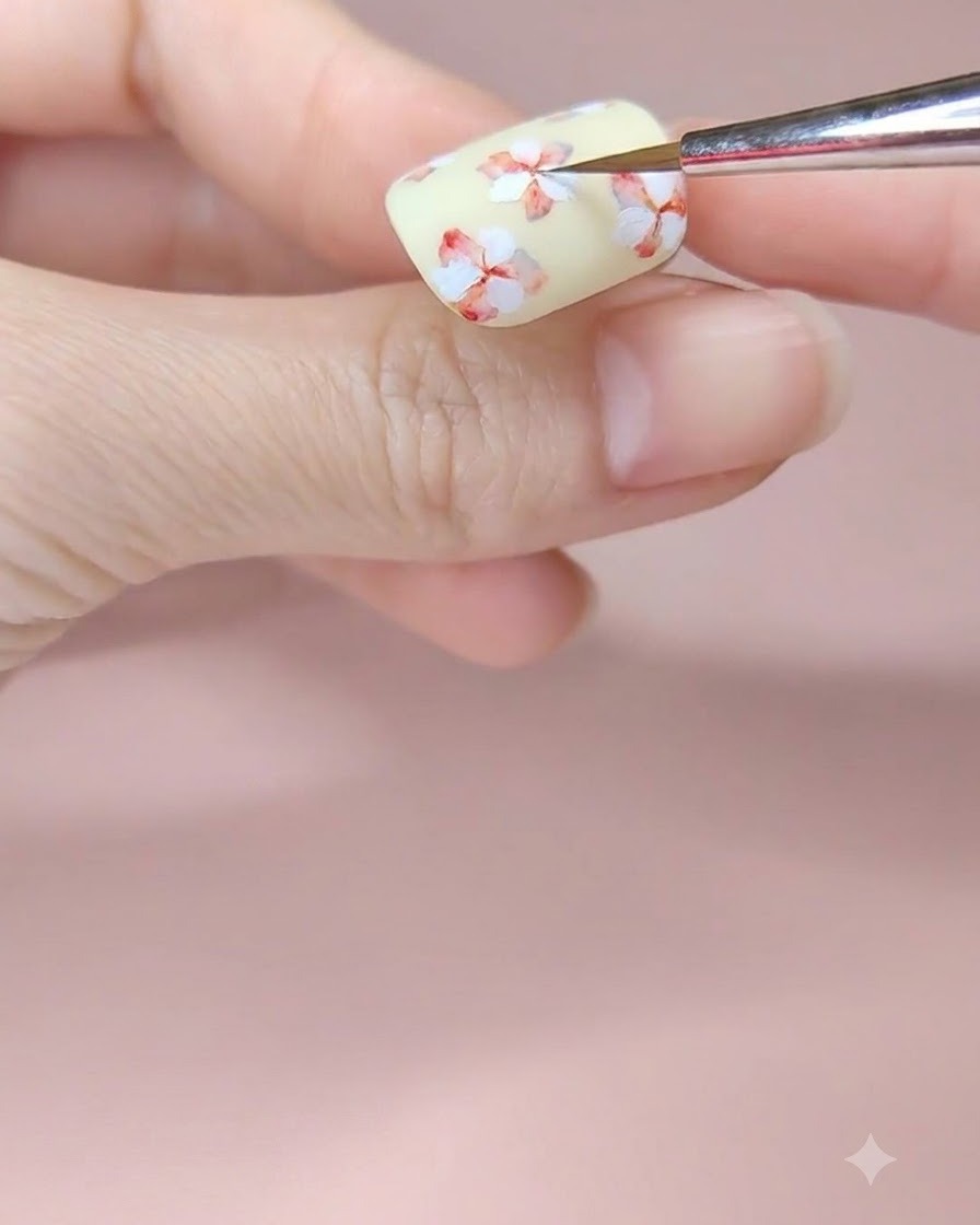 Ein feiner Nailart-Pinsel fügt der Mitte einer weiß gemalten Blume auf einem blassgelben Nagel einen Hauch von rotem Aquarellpigment hinzu.