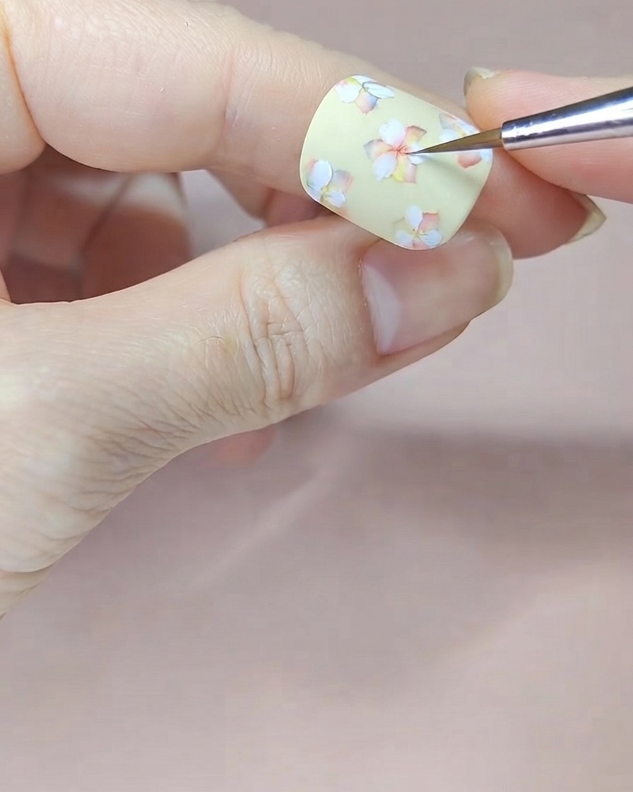 Ein feiner Pinsel trägt rote Aquarellfarbe auf die Mitten der weißen Blumen auf einem blassgelben Nagel auf.