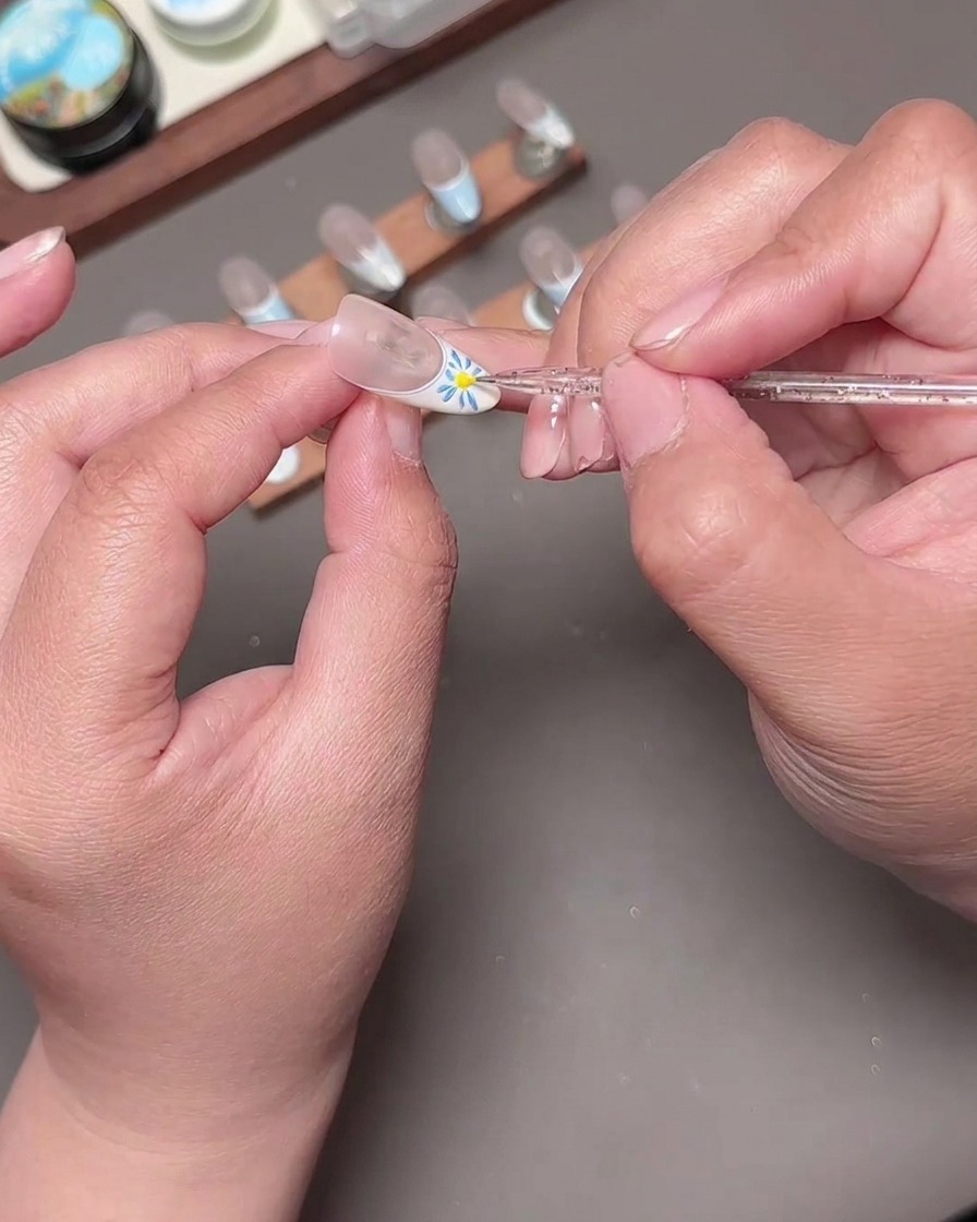 Ein Dotting-Tool platziert einen leuchtend gelben Mittelpunkt in eine blau gemalte Blume auf einem French-Maniküre-Nagel.
