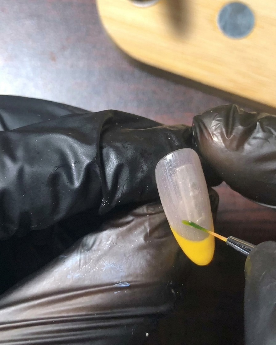 Ein feiner Detailpinsel malt ein winziges grünes Stiel- und Blattdetail nahe der Smile-Line des Nagels.