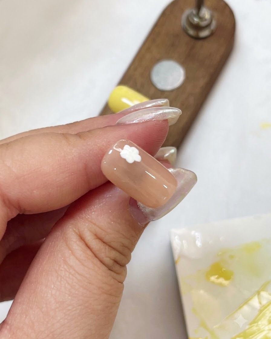 Nahaufnahme eines Nude-Nnagels mit einem weißen Blumenmuster, das mit einer kleinen violetten Stiftlampe ausgehärtet wird.