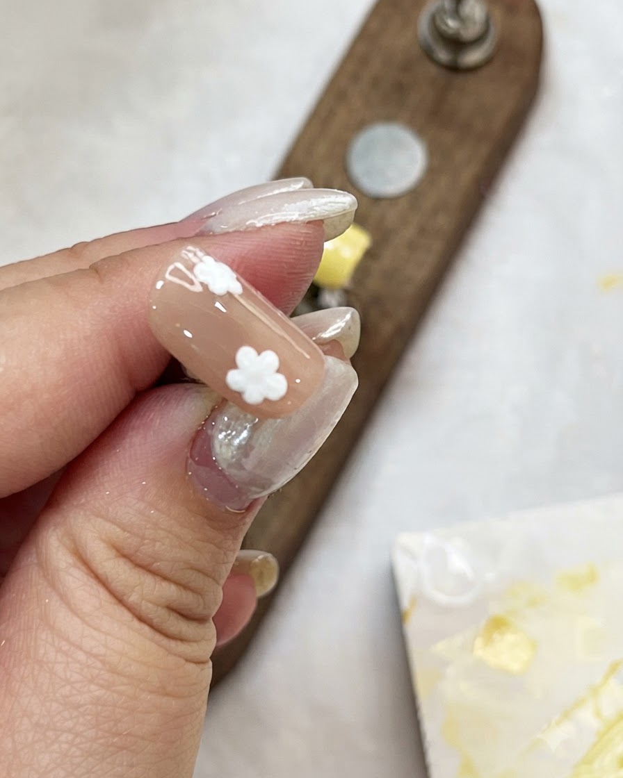 Eine Hand hält eine Nude-farbene Nagelspitze und fügt weiße Punktblüten hinzu, um Blumen zu kreieren.