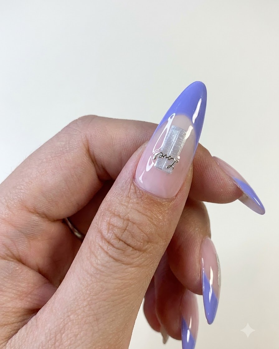 Eine Nahaufnahme eines Daumennagels mit blauem French Tip, silbernem Folienelement als Basis und einem zarten Buchstaben-Sticker.