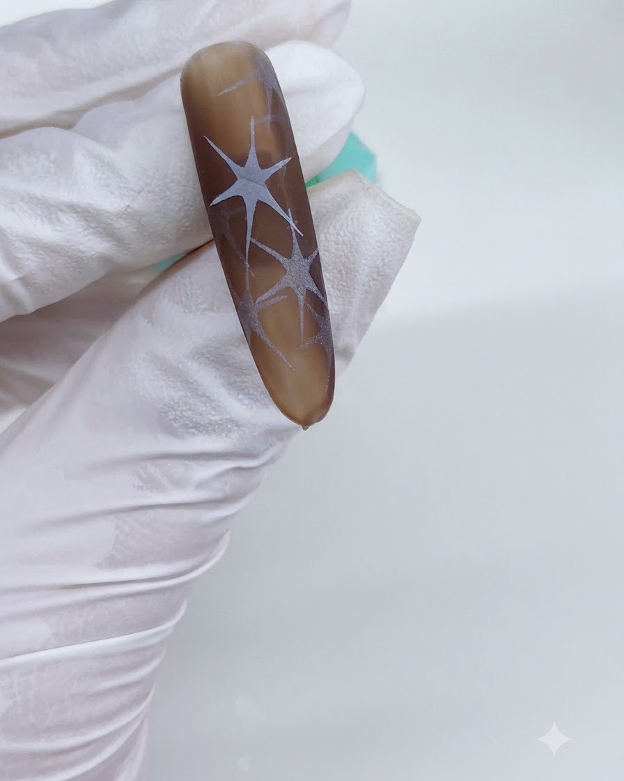 Eine behandschuhte Hand hält einen braunen Nagel unter eine UV-Lampe, wobei ausgehärtete weiße Sternmuster sichtbar sind.