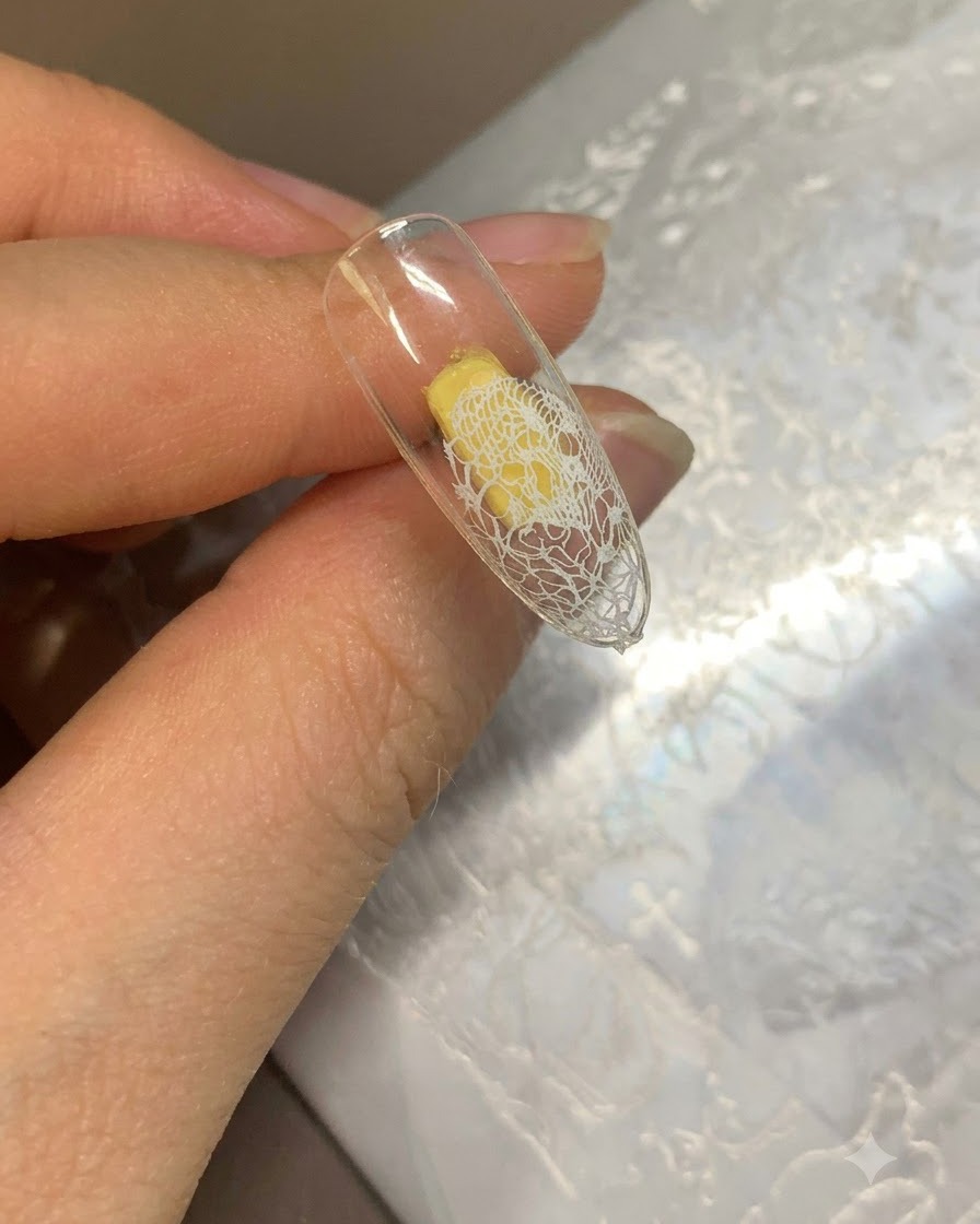Eine klare Nagelspitze mit einem weißen Spitzenmuster am Ende und einer beigen Gel-Basis auf dem restlichen Nagel.