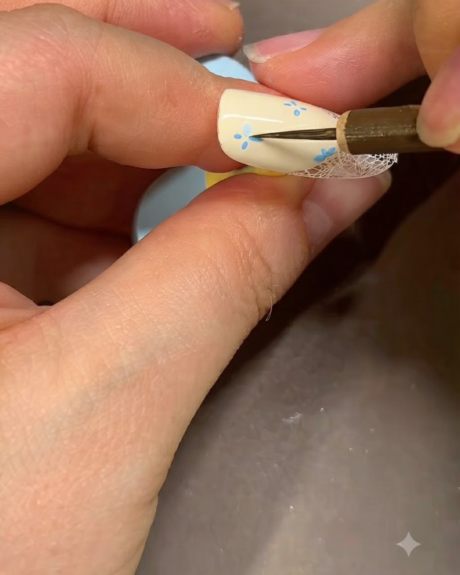 Ein feiner Nail-Art-Pinsel fügt detaillierte blaue Akzente in der Mitte einer gemalten Blume auf einem beigen Nagel hinzu.