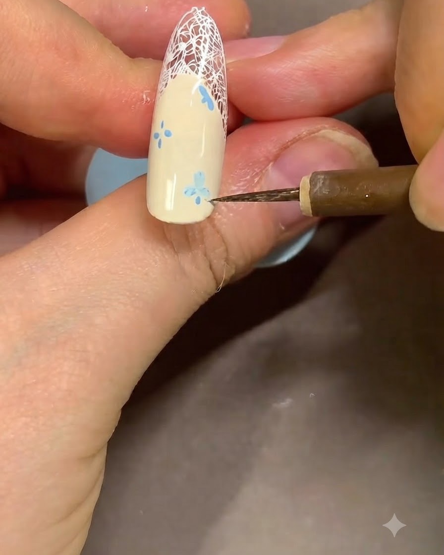 Nahaufnahme eines Nail-Artists, der mit einem feinen Detail-Pinsel die Kanten eines blauen Blumen-Designs nahe der French-Spitze säubert.