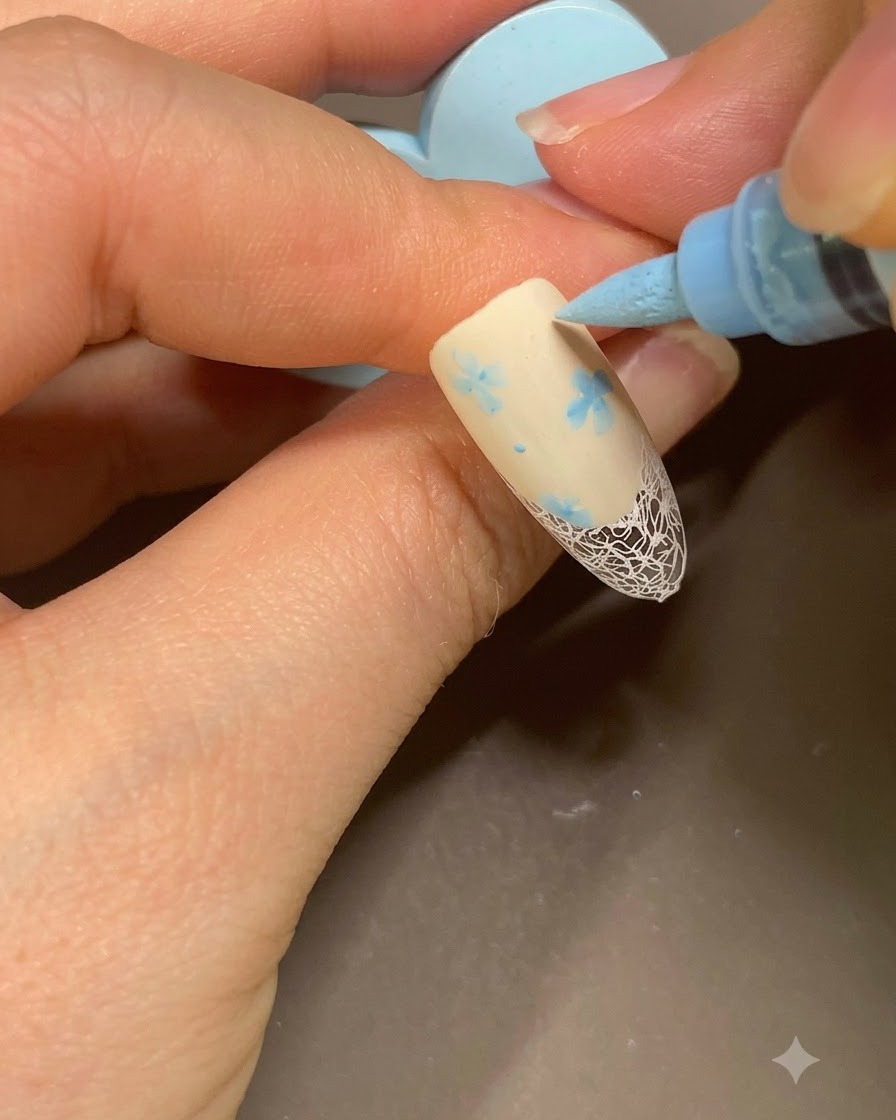 Ein blauer Nail-Art-Marker fügt dem floralen Muster auf einer beigen French-Maniküre letzte kleine Punkte hinzu.
