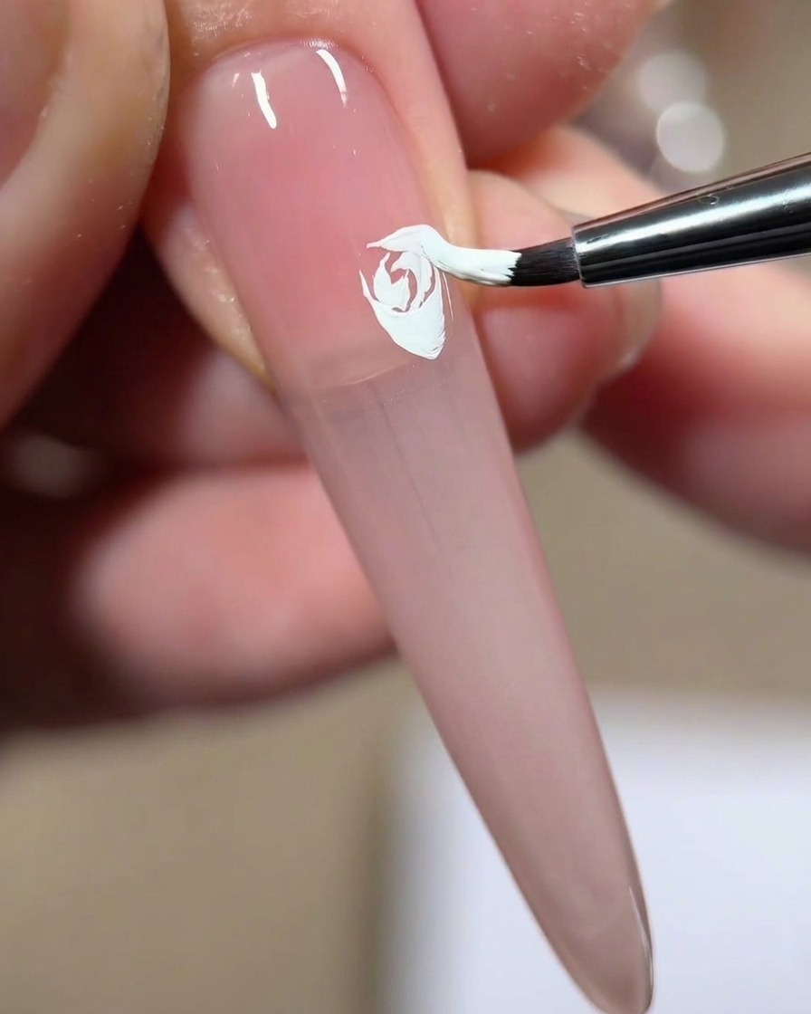 Ein Nagelpinsel drückt und zieht weißes Gel, um einer gemalten weißen Rose auf einem rosa Nagel ein viertes geschichtetes Blütenblatt hinzuzufügen.