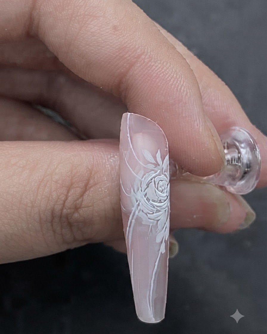 Ein Nahbild, das zeigt, wie ein hellrosa Farbton mit einem Pinsel auf die Mitte eines dreidimensionalen weißen Rosen-Nail-Designs aufgetragen wird.