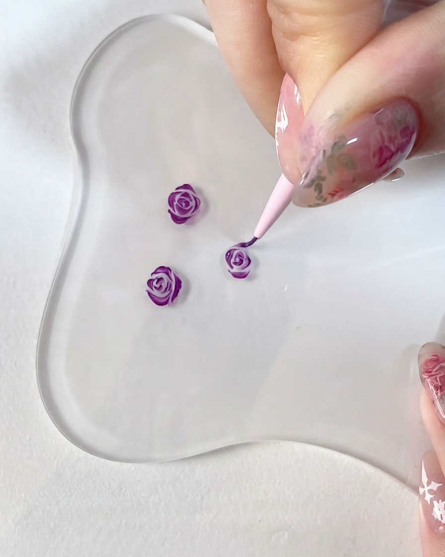 Eine Nahaufnahme einer Hand, die einen Feindetailpinsel hält und akribisch dunkelviolette Akzente auf ein kleines 3D-Rosendesign auf einer klaren Glaspalette malt.