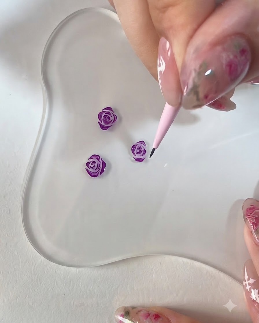 Eine Nahaufnahme einer Hand, die einen rosa Detailpinsel verwendet, um die Kanten einer 3D-gemalten lila Rose auf einer klaren Palette zu verfeinern.