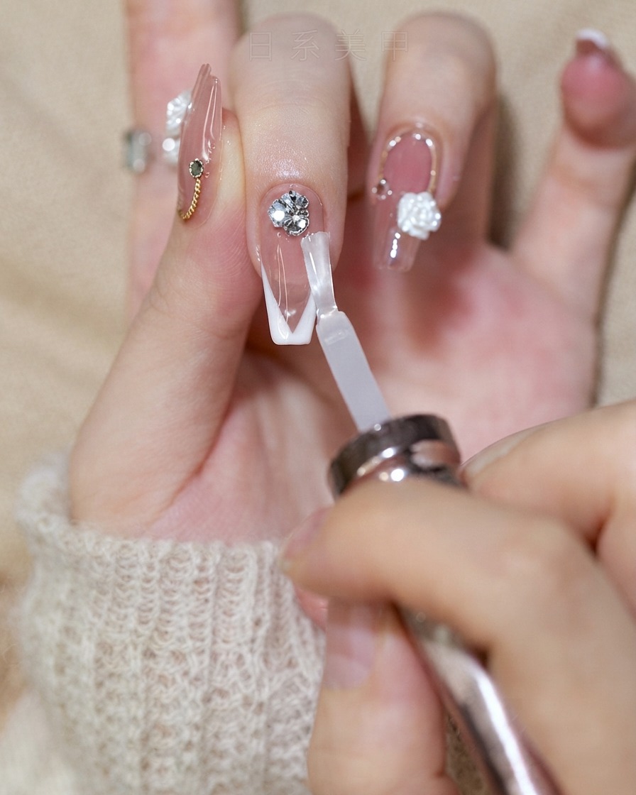 Ein Nagelpinsel trägt einen glänzenden Top Coat auf einen French-Tip-Nagel auf und spart dabei das Strassstein-Cluster aus.