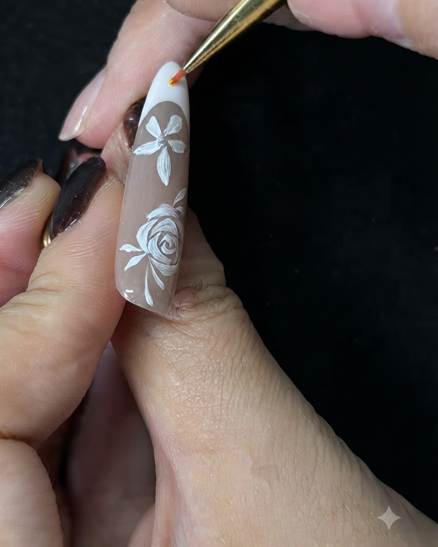 Nahaufnahme einer Hand, die mit einem feinen Detailpinsel kleine weiße Akzentranken um eine handgemalte weiße Rose auf einem Nude-Gelnagel malt.