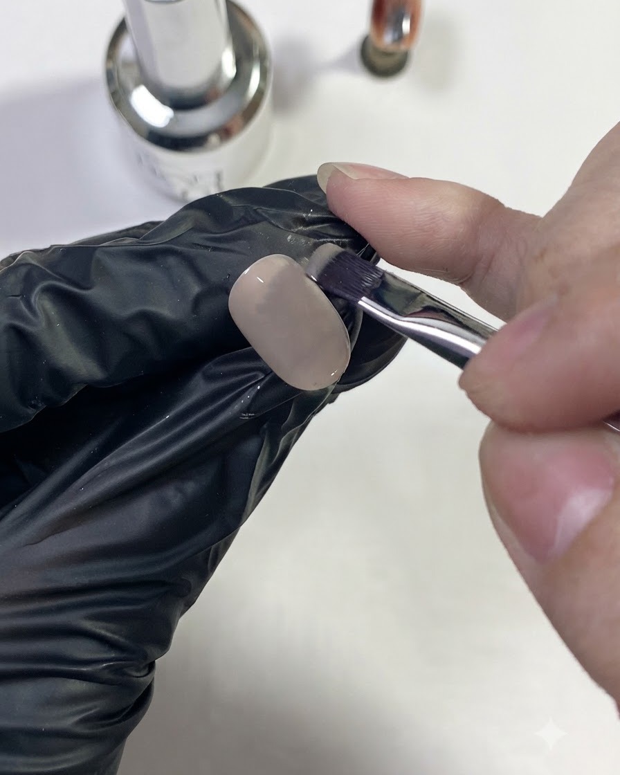 Eine behandschuhte Hand trägt eine Nude-Basisfarbe auf eine Nagelspitze auf.