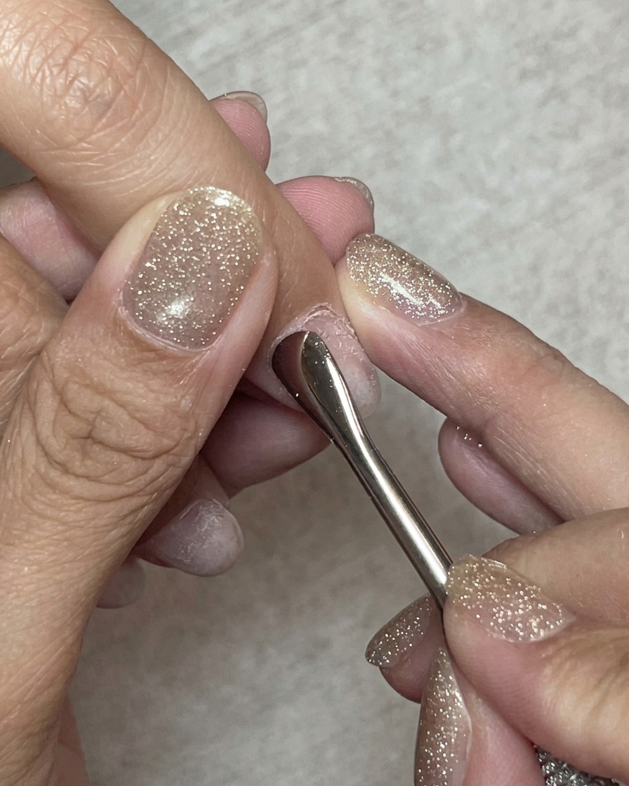 Nahaufnahme eines Nagelhautschiebers aus Stahl, der die Nagelhaut an einem Naturnagel vorsichtig zurückschiebt.