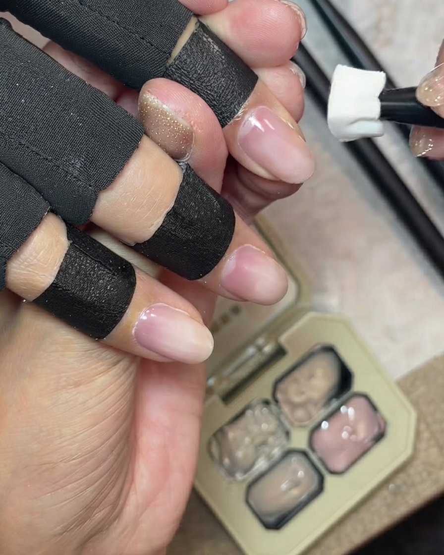 Ein Schwämmchen, das milchig-nuedes Covergel waagerecht auf die natürliche Nagelspitze eines Fingers auftupft, mit einer Gelpalette im Hintergrund.