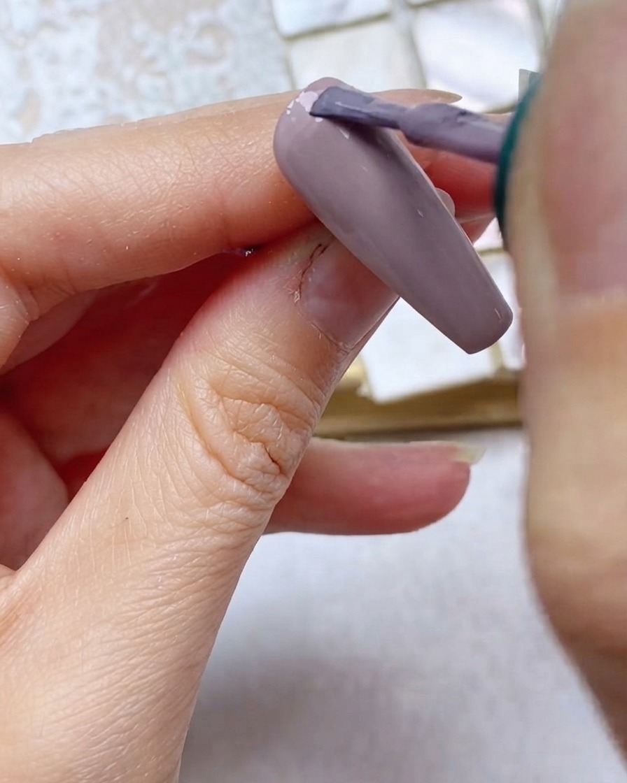 Ein Pinsel trägt graulila Gelpolitur auf das obere Drittel des Nagels nahe der Nagelhaut auf.