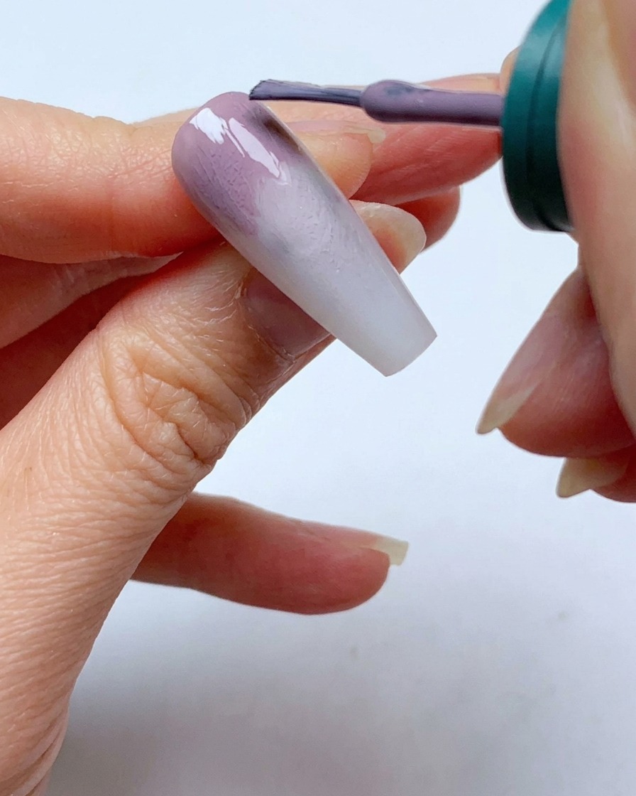 Ein Nagellackpinsel trägt eine zweite Schicht graulila Gel nahe der Nagelhaut eines Nagels auf.