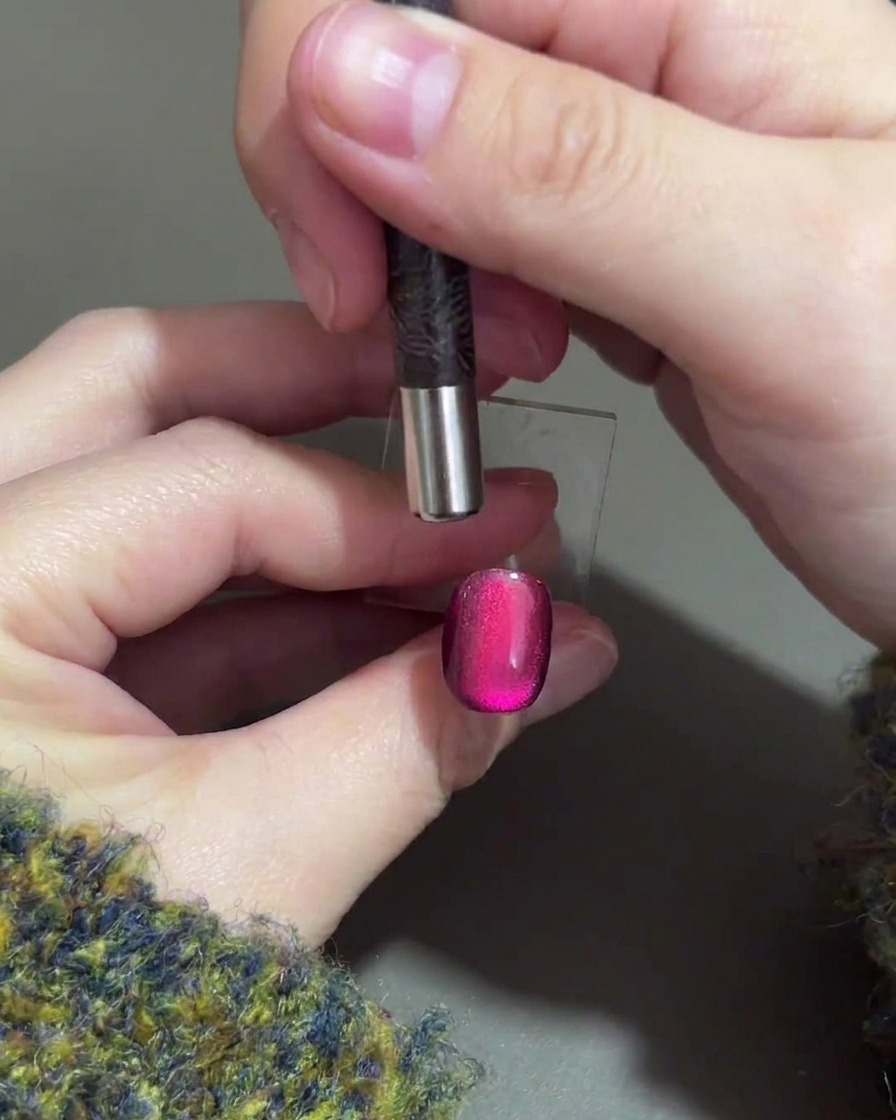 Eine Hand hält einen zylindrischen Magneten über die obere Kante eines violetten Gelnagels, um das magnetische Highlight nach unten zu schieben.