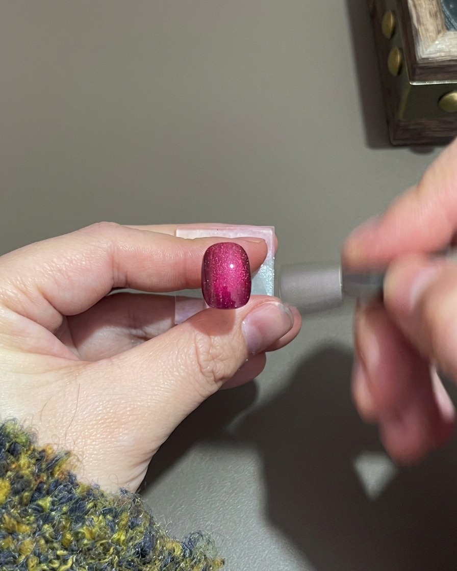 Eine Hand hält einen kleinen zylindrischen Magneten in der Nähe eines violetten Cat-Eye-Gelnagels.