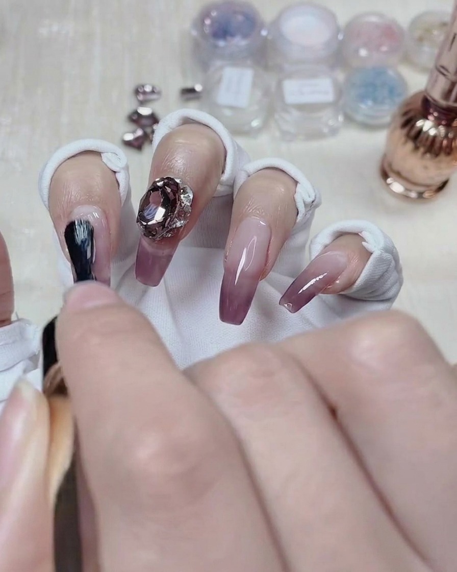 Ein Nail Artist trägt Top Coat um die Basis eines Kristalls auf einem fertigen Gradient-Nagel auf.