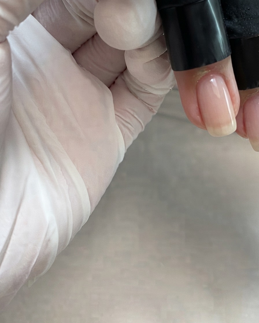 Ein Flaschenpinsel trägt ein transparentes, nudefarbenes Pink-Gel in der Mitte eines Fingernagels auf.