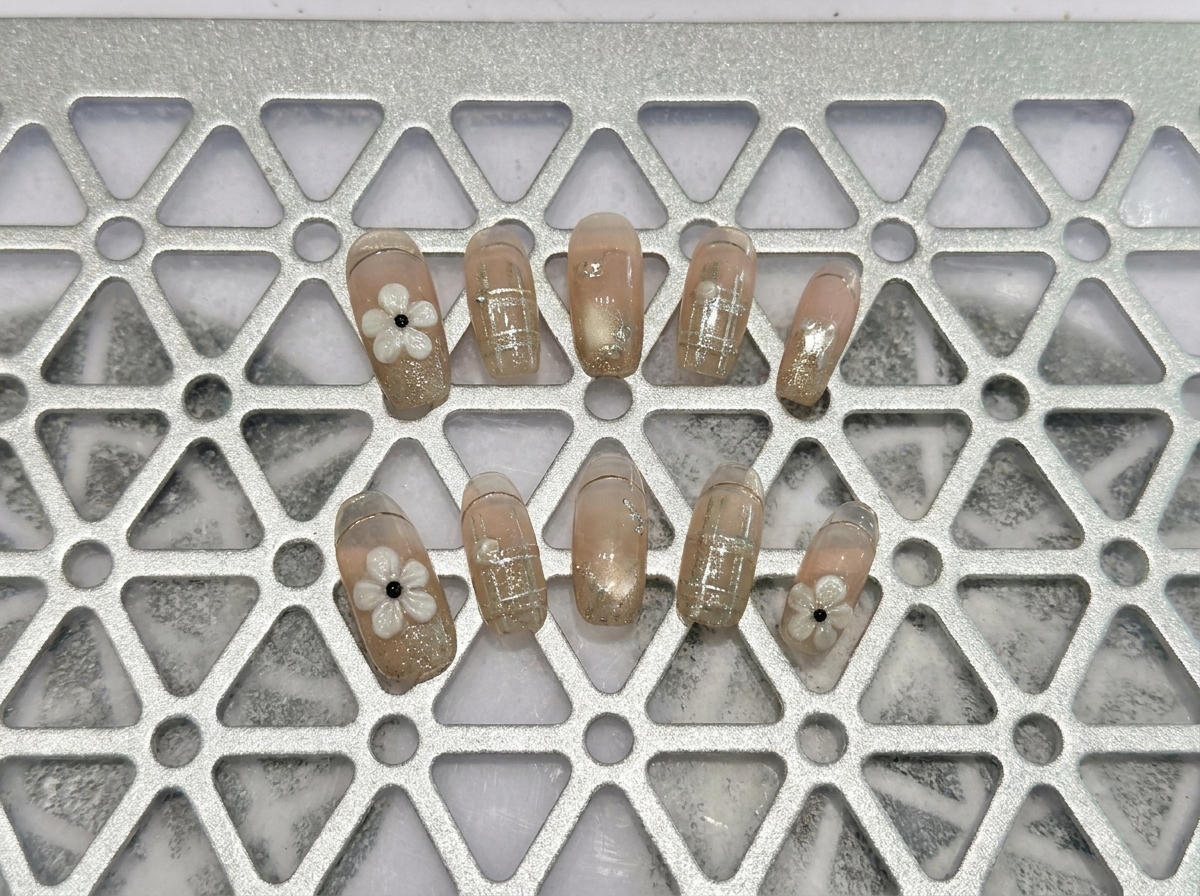 Eine Nahaufnahme einer Hand, die einen strukturierten weißen Blumen-Charm und einen feinen Glitzerverlauf auf einer neutralen Gelbasis zeigt.