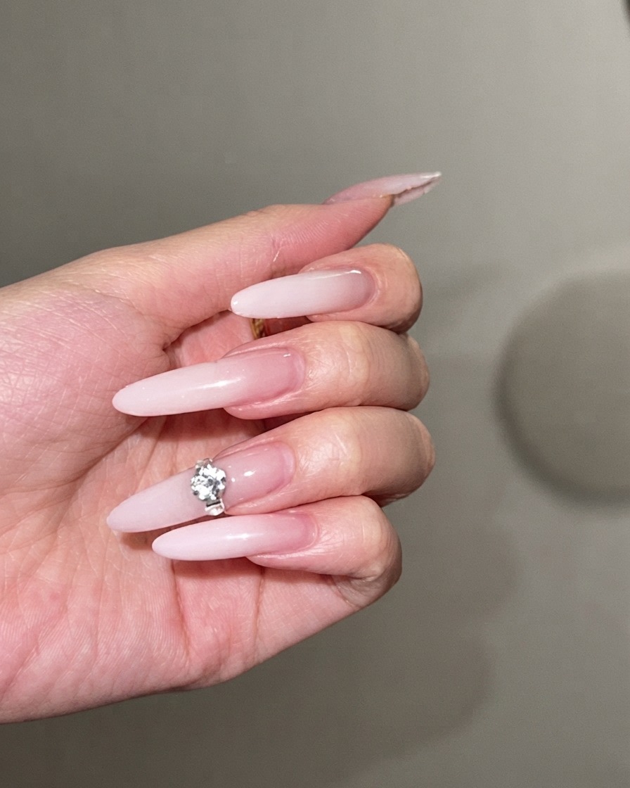 Ein großer, funkelnder Strassstein wird in die Mitte eines langen Stiletto-Nagels mit milchweißem Ombré gedrückt.