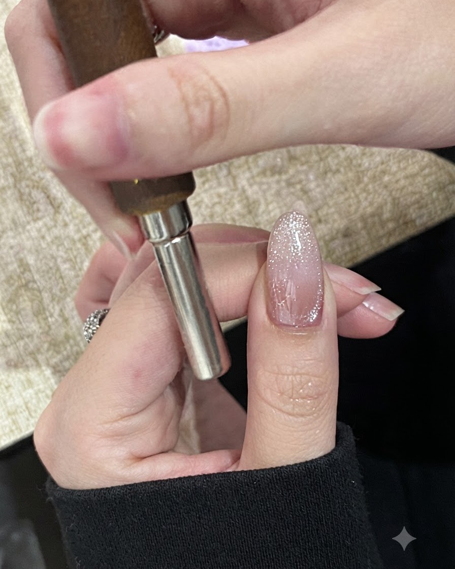 Eine Hand hält einen zylindrischen Magneten waagerecht über die Spitze eines glitzernden, noch nicht ausgehärteten Cat-Eye-Gel-Nagels.