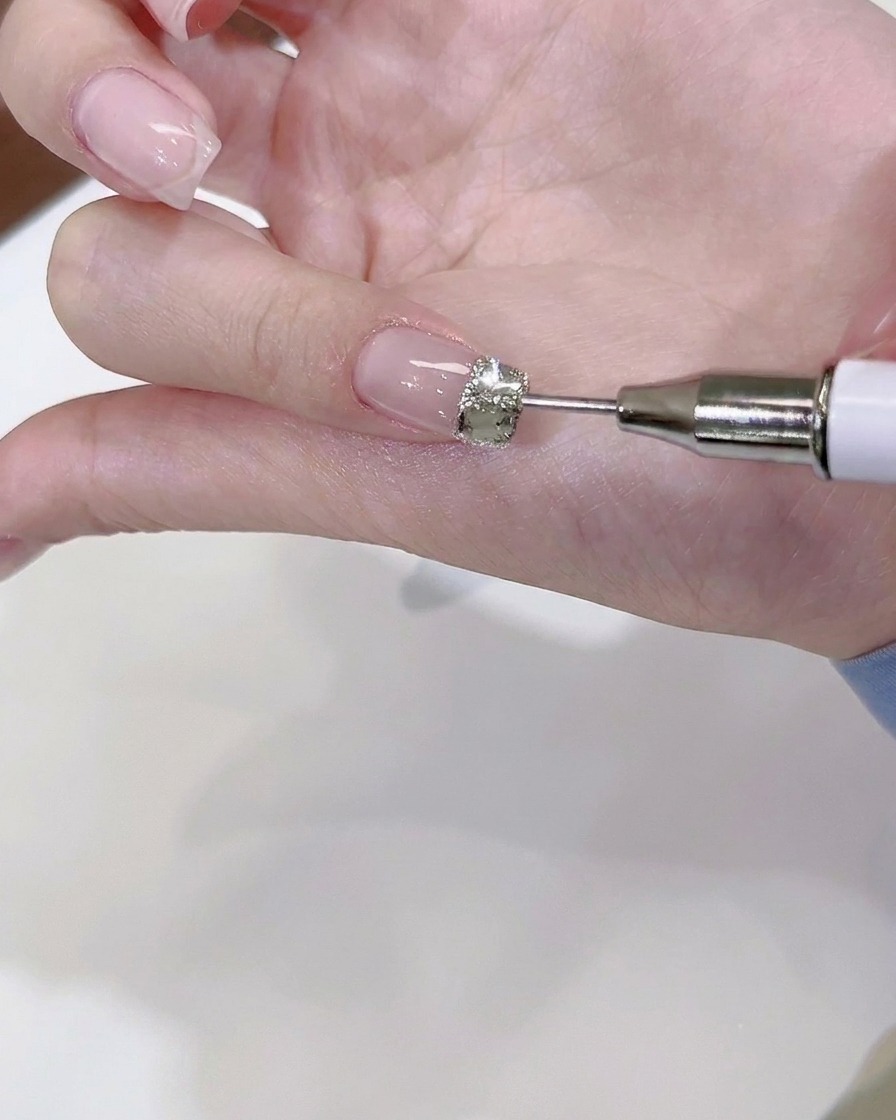 Ein feines Dotting-Tool platziert winzige Stahlperlen und Mini-Kristalle um einen großen zentralen Strassstein auf einem Fingernagel.