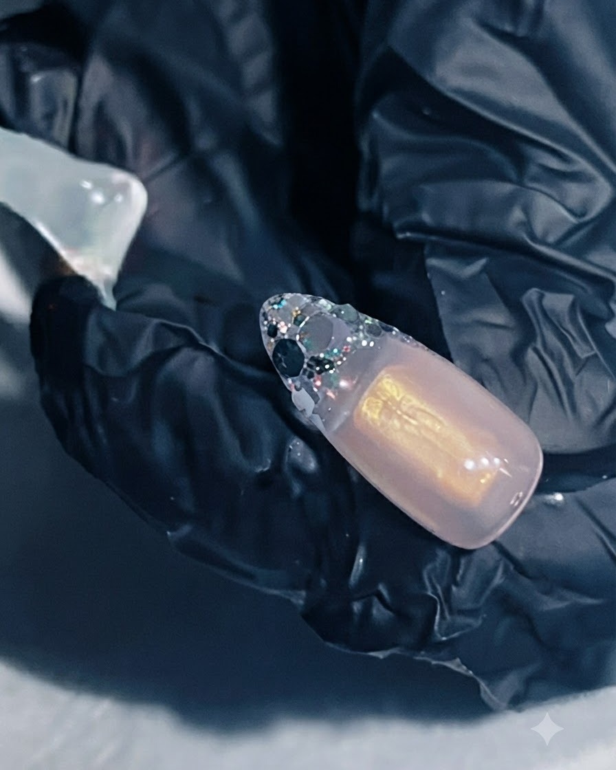Eine Hand hält eine Nagelverlängerung mit aufgetragenem Verstärkungsgel über dem Glitter-French-Bereich.
