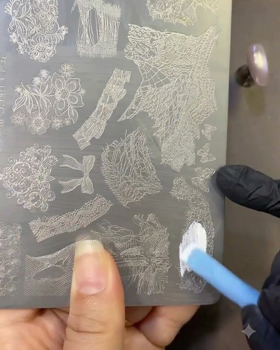 Ein Pinsel trägt weißen Stamping-Lack auf eine Metallplatte mit eingravierten Spitzenmustern auf.