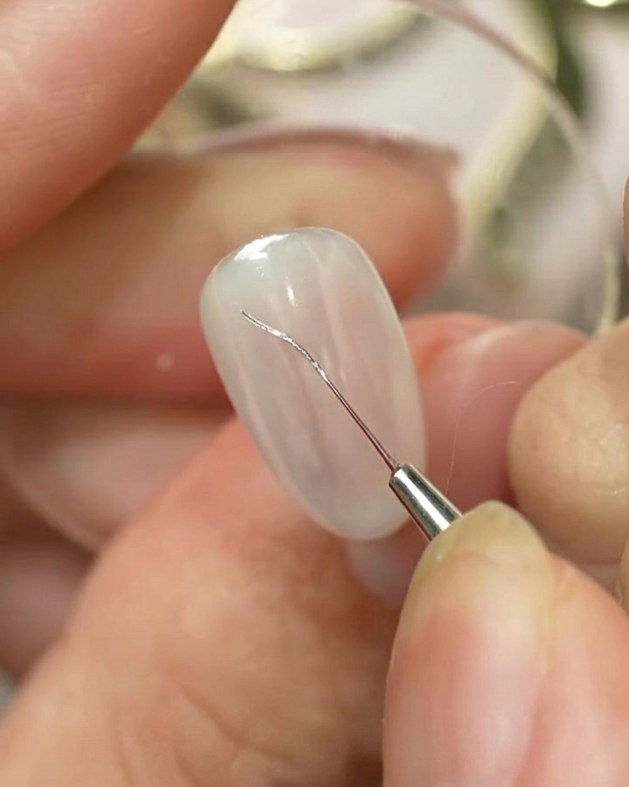 Ein feiner Nail-Art-Liner-Pinsel zeichnet eine silberne Schleifenkontur auf einen hellfarbigen, manikürten Nagel.