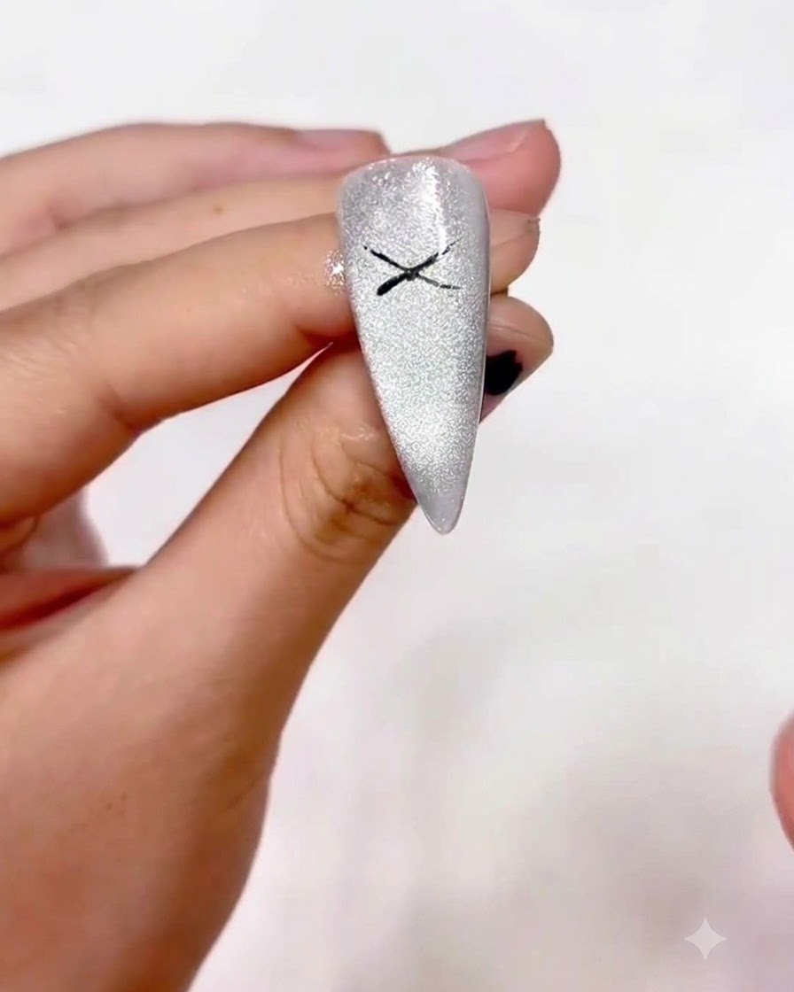 Nahaufnahme einer Hand mit einem feinen Liner-Pinsel, die ein schwarzes X in die Mitte eines schimmernden silbernen Cat-Eye-Nagels zeichnet.