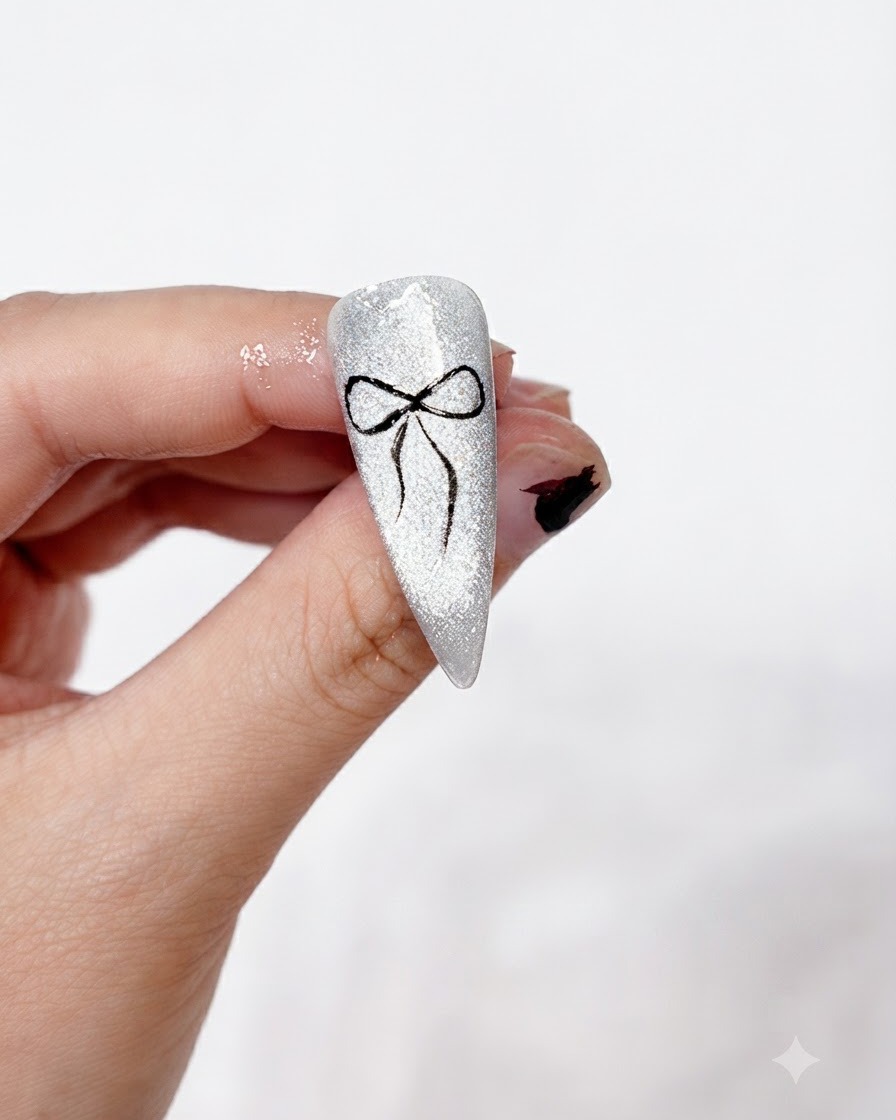 Ein fertiges, sauber gemaltes schwarzes Schleifen-Nail-Art-Design auf einem glitzernden silbernen Cat-Eye-Hintergrund, gehalten zwischen den Fingern.