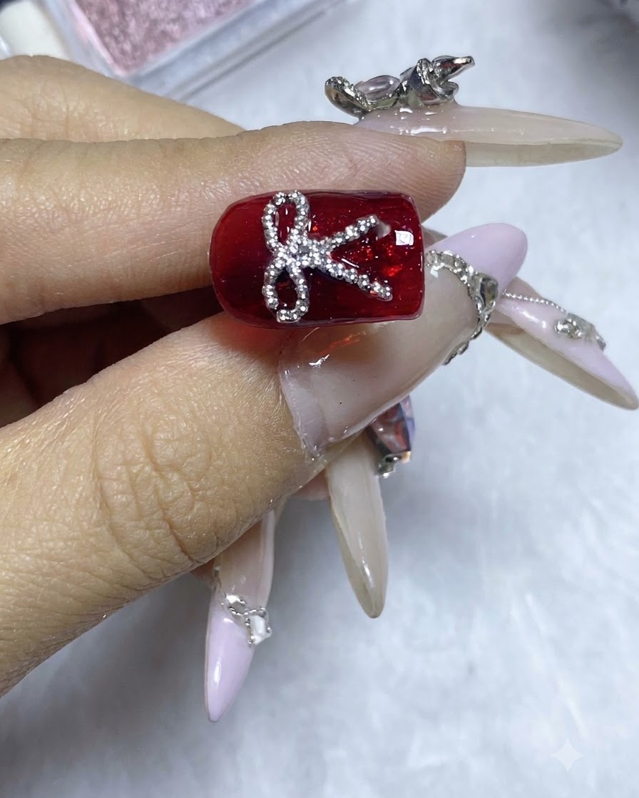 Ein fertiges rotes Nageldesign mit einer glitzernden Stahlperlen-Schleife unter hellem Studiolicht.