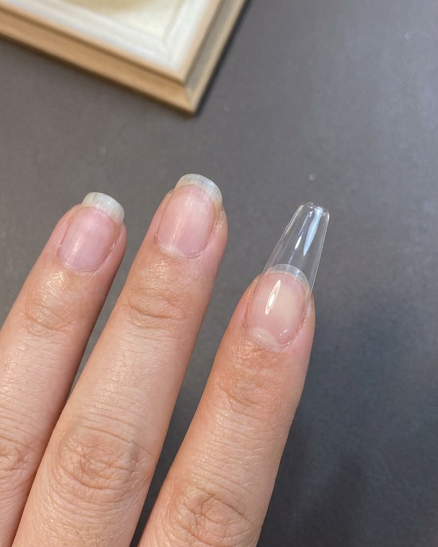 Nahaufnahme einer Hand mit einem Gelpinsel, die eine erste Schicht transluzenter Nude-Basisfarbe auf eine verlängerte Nagelspitze aufträgt.
