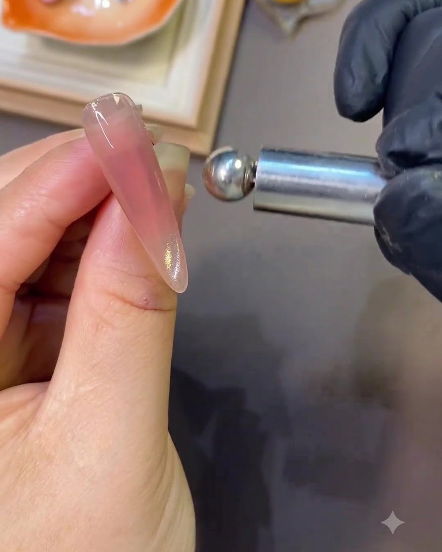 Ein Magnetwerkzeug wird nah an einen glänzenden rosa Nagel gehalten, um eine seitliche Lichtreflexion des Cat-Eye-Effekts zu erzeugen.