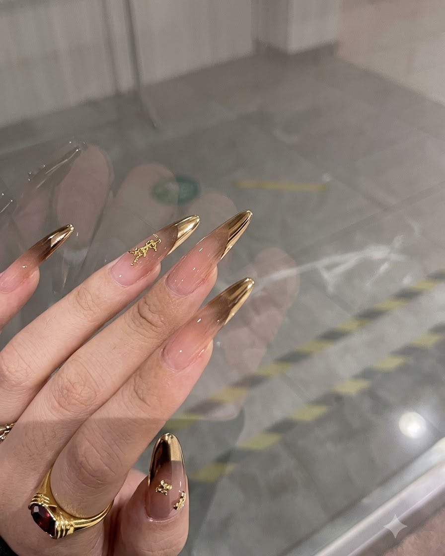 Eine Hand zeigt lange Mandel-Nägel mit Bronze-Chrome-French-Spitzen und Goldakzenten.