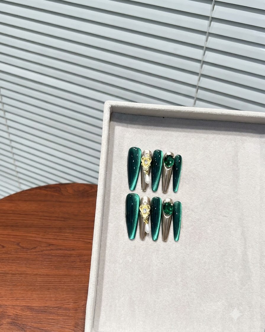 Eine schräge Nahaufnahme einer Aufbewahrungsbox mit einem vollständigen Set aus tiefgrünen, mandelförmigen Press-on Nägeln mit Metallic- und Strassakzenten.